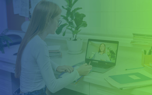 A student sitting at a desk with a notebook and pen, looking at a laptop screen showing a teacher on a video call, with books and a potted plant nearby.